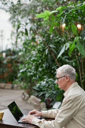 An older man with short grey hair sits using his computer in a garden setting