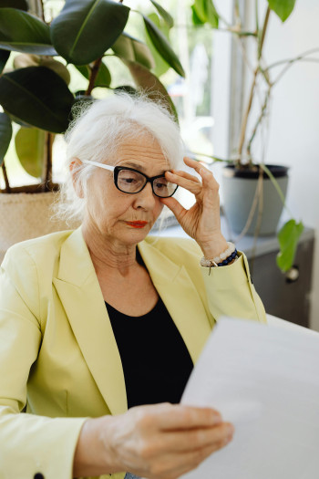 An older woman in a yellow blazer sits looking at a report. She is adjusting her glasses.