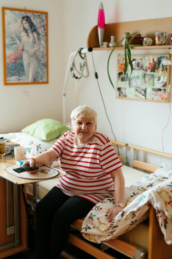 An older woman sits in a hospital-style bed in a cosy room. She has a TV remote in her hand. She has short white hair and is wearing a striped red and white t-shirt and black pants.