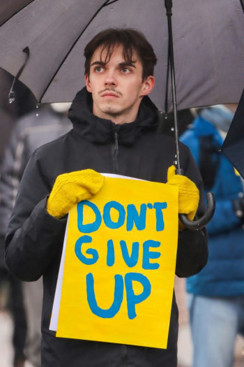 A young man stands outside holding an umbrella and a bright yellow sign that says don't give up