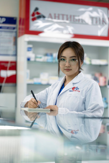 A woman with long dark hair who wears glasses stands behind a tall desk taking notes. She has a white coat on and is in a pharmacy.