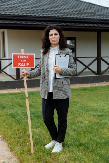 A dark haired woman stands outside a house holding a for sale sign and a clipboard.