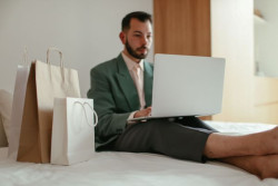 A man in a suit sits on a bed using a laptop. He is surrounded by shopping bags.