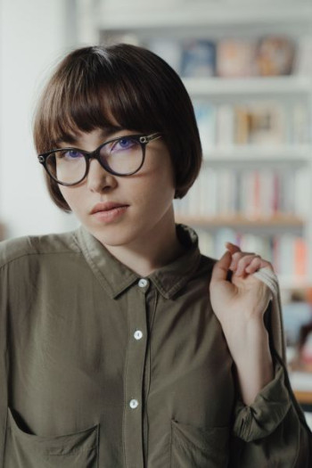A white woman with a dark brown bob haircut stands looking at the camera. She is wearing smart glasses and an olive shirt.