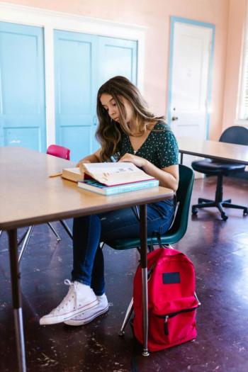 A student sits at a desk looking at a text book. Her red bag sits on the floor beside her.