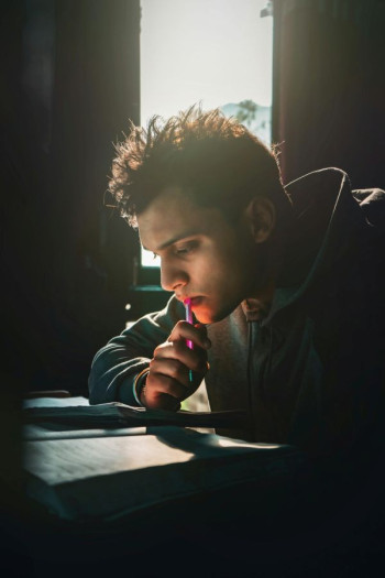 A male student in a hoody sits staring at a paper on a desk. He is holding a pen.