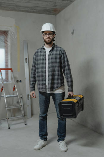 A tradie standing inside a house. He is wearing a white hard hat, checked shirt, jeans, and is carrying a tool box.