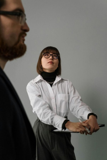 A woman with a dark bob haircut and glasses stands looking up. She's holding a clipboard and is wearing a white shirt and dark pants. A man stands closer to the camera but we can't see who it is.