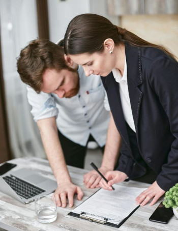 Two white people - a man and a woman - in their thirties stand over a desk looking at a clipboard.