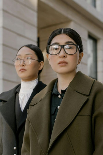 Two woman with long dark hair pulled back stand looking at the camera. They both have glasses and fair skin. They are dressed professionally.