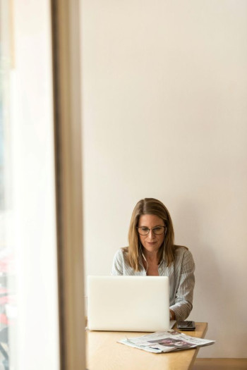 A woman with long blonde hair and glasses sits at a desk behind a laptop.