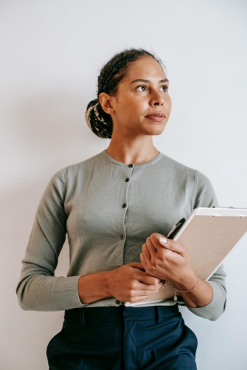 A brown woman with tied back hair stands looking to the left holding a clipboard. She looks contemplative. She is wearing a green buttoned up cardigan and dark blue pants.