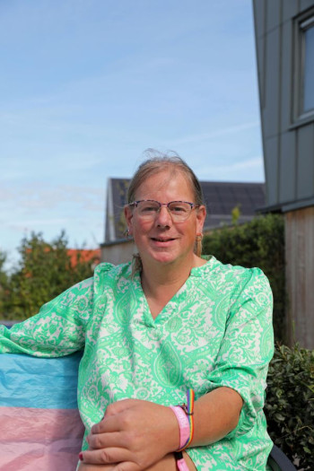 A white woman with her hair pulled back sits on a chair looking at the camera. She has a green and white patterned top and is wearing a rainbow bracelet. 