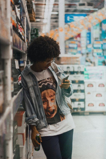 A person with an afro stands in a store leaning against the display. They are wearing a Home Alone t-shirt, jeans, and denim jacket. They have glasses and are looking at their phone.
