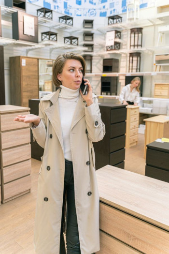 A woman in a beige trench coat stands in a modern furniture store and is on the phone.