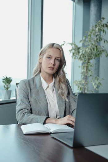 A young blonde woman wearing a grey suit sits at a desk behind a laptop.