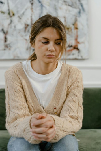 A woman sits looking glum. She is wearing a white tshirt, beige cardigan, and jeans.