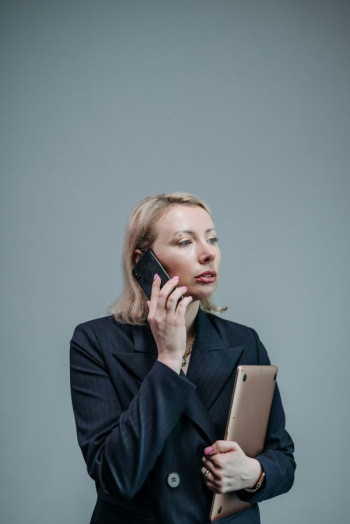 A white woman with blonde hair stands holding a laptop and speaking on the phone