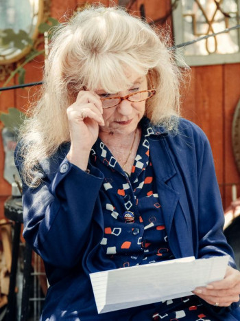 An older white woman with long blonde hair stands reading a letter. She is wearing navy clothes and glasses.
