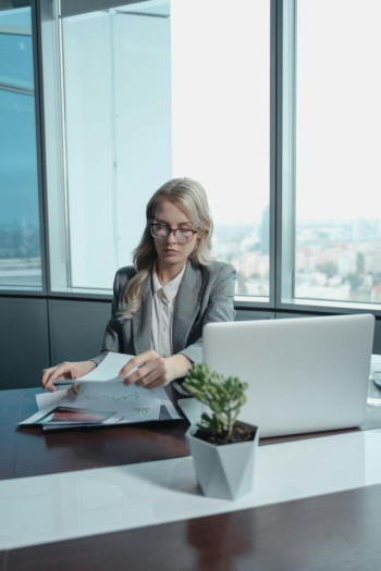 A woman with blonde hair and glasses sits in a corporate office with paperwork and a laptop.