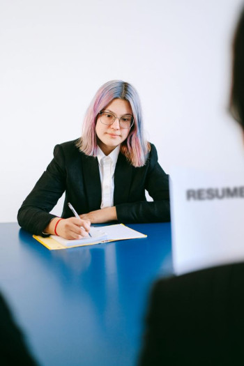 A woman in business dress with pink hair sits at a table writing on a document.