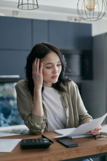 A woman with long dark hair sits at a desk looking at a piece of paper. A calculator sits on the desk too.