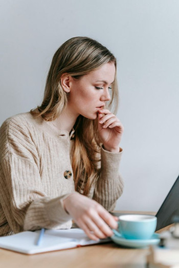 A woman with long blonde hair sits at a desk with an open laptop and a coffee.