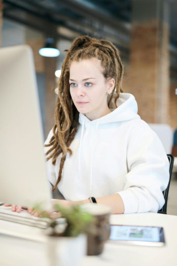 A white woman in a white hoody sits at a computer. She has dreadlocks.