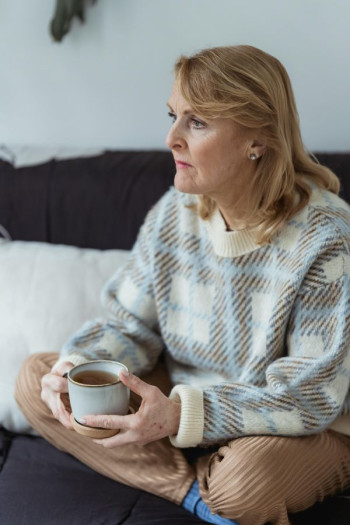 A woman in a warm top and beige pants sits inside looking into the distance. She is holding a cup of tea.
