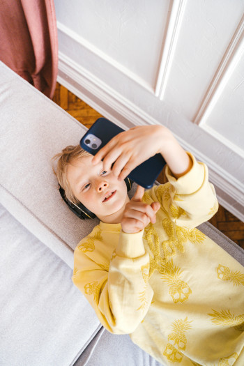A blonde child lies on their back on a sofa, holding a cell phone above their face.
