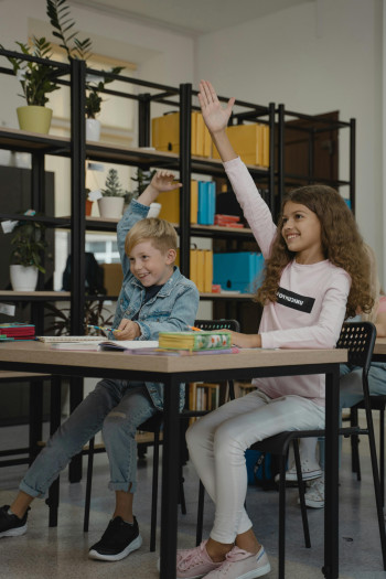 Two children sit at a desk with their hands raised.