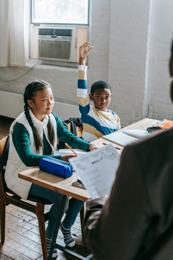 Two children sit at a school desk with books and papers in front of them. One of them faces the teacher with his hand raised.