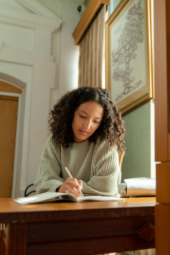 A woman with dark curly hair leans on a desk while writing in a notebook.