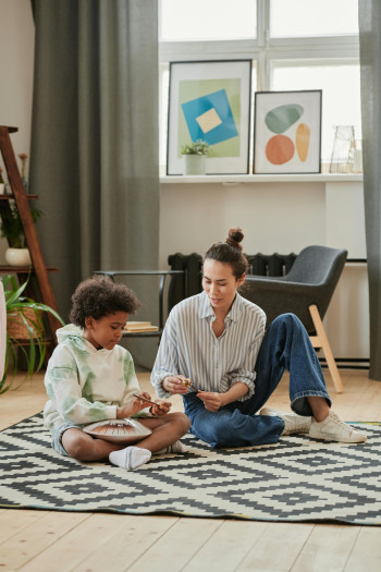 A woman and a child sit on a rug on the floor and play with a steel drum.