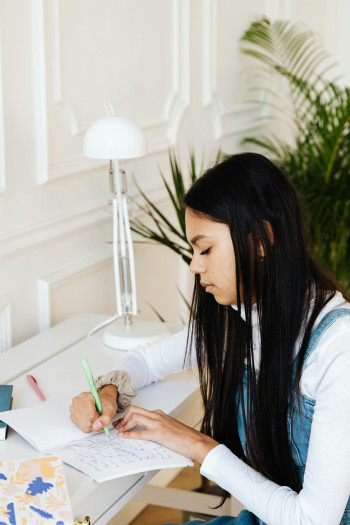 A student with long, dark hair sits at a desk and writes in a notebook.