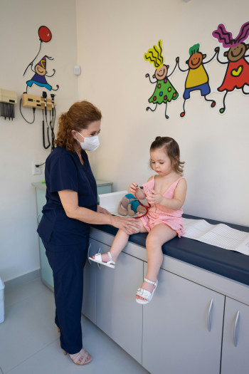 A doctor or nurse in scrubs lets a young child play with a stethoscope during a routine examination.