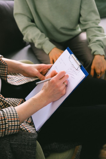 Two people sit opposite each other, one is taking notes on a clipboard.