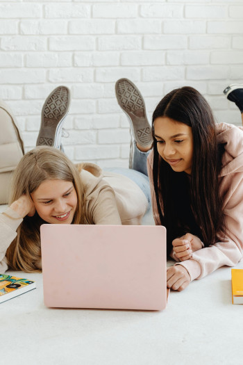 Two girls lie on their fronts on the ground, using a laptop.