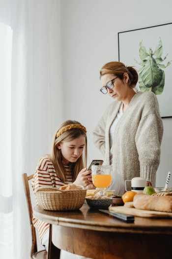 A girl sits at a table and looks at her cell phone. Her mother stands over her, with a hand on her hip.