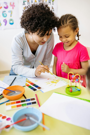 A teacher and a young child lean over a table to paint a picture. The table is covered with paint, paper, and paintbrushes.