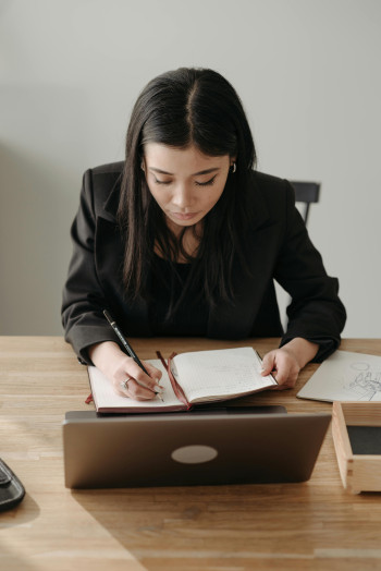 A woman sits at a desk with her laptop and notebook in front of her. She is writing in the notebook.