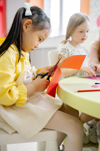Young children sit at a colourful table and use scissors to cut shapes out of coloured paper.
