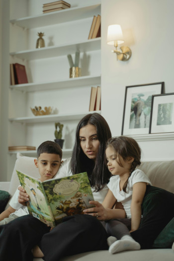 A woman sits on a couch reading to two children, who sit on either side of her.