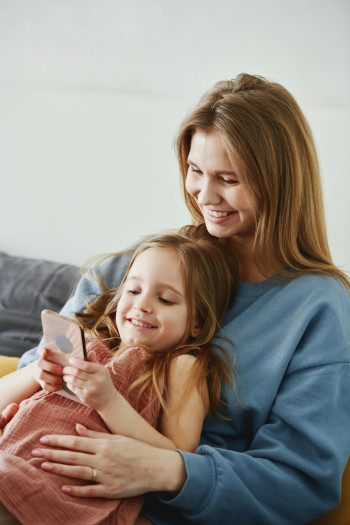 A woman sits on a couch with a young girl sitting in her lap, as they both look at a cell phone screen.