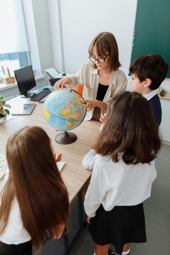 A teacher sitting at a desk points at a globe while three children gather around and look.