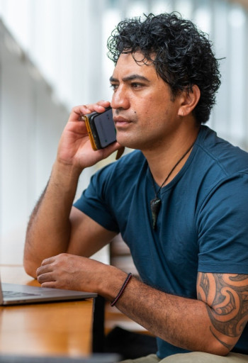 A Māori man sits at a table or counter, holding his cellphone to his ear.