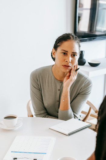 A woman sits at a table with a notebook in front of her. She holds a pen in her hand and looks at someone across the table she is talking to.