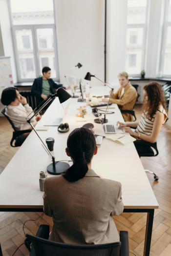 Five people sit at a table in an office having a meeting. Some have laptops in front of them.