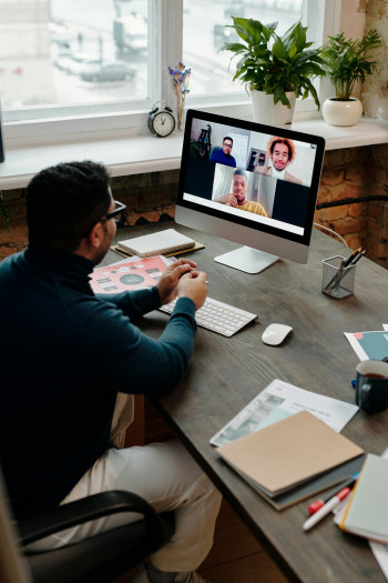 A man wearing glasses sits at his desk with three other people on his computer screen, in a video meeting.