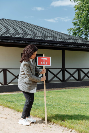 A woman stands on the path outside a house and plants a for sale sign in the dirt.